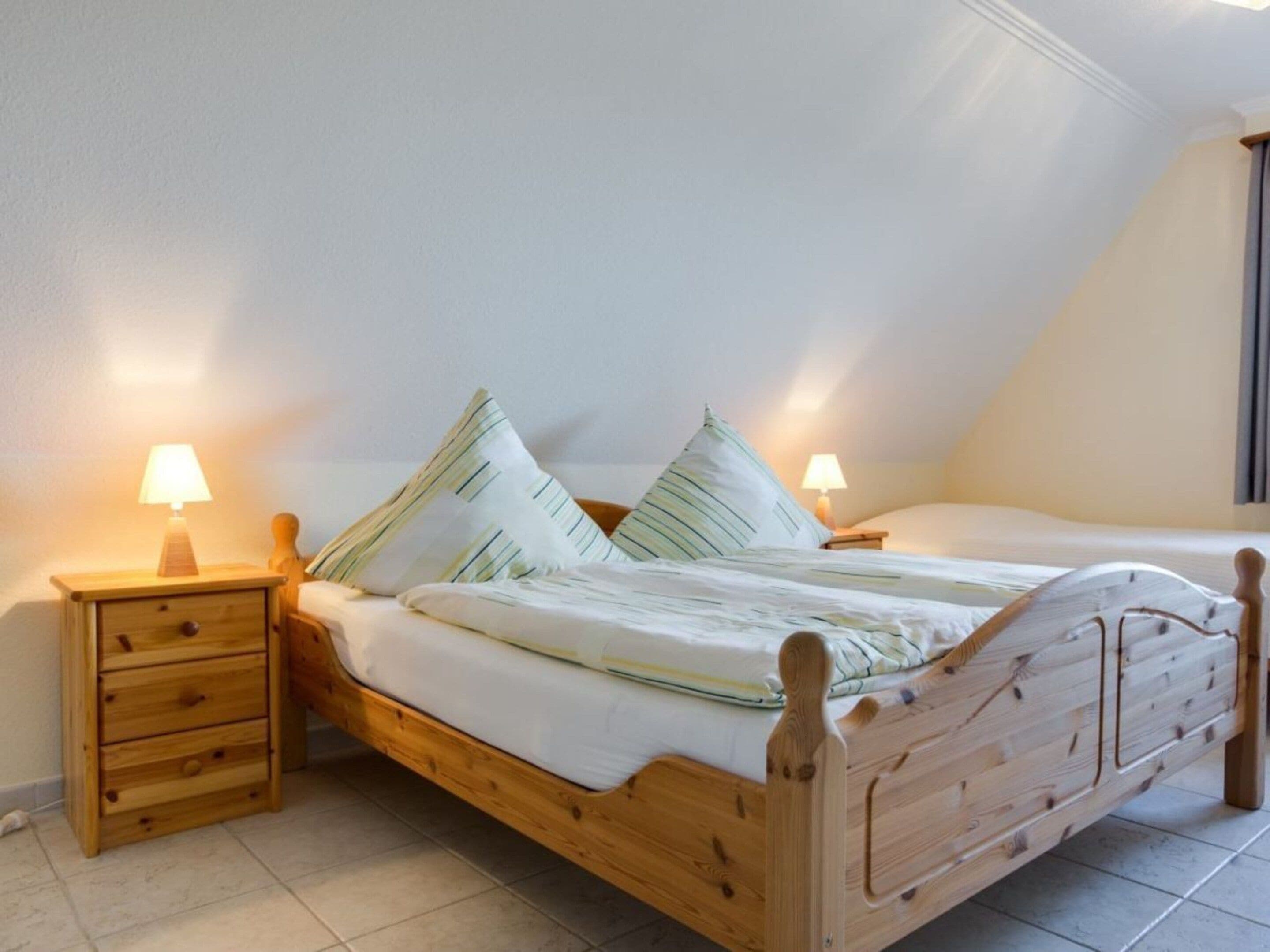 A wooden bed with two nightstands, each with a lamp, in a room with white walls and a sloped ceiling.