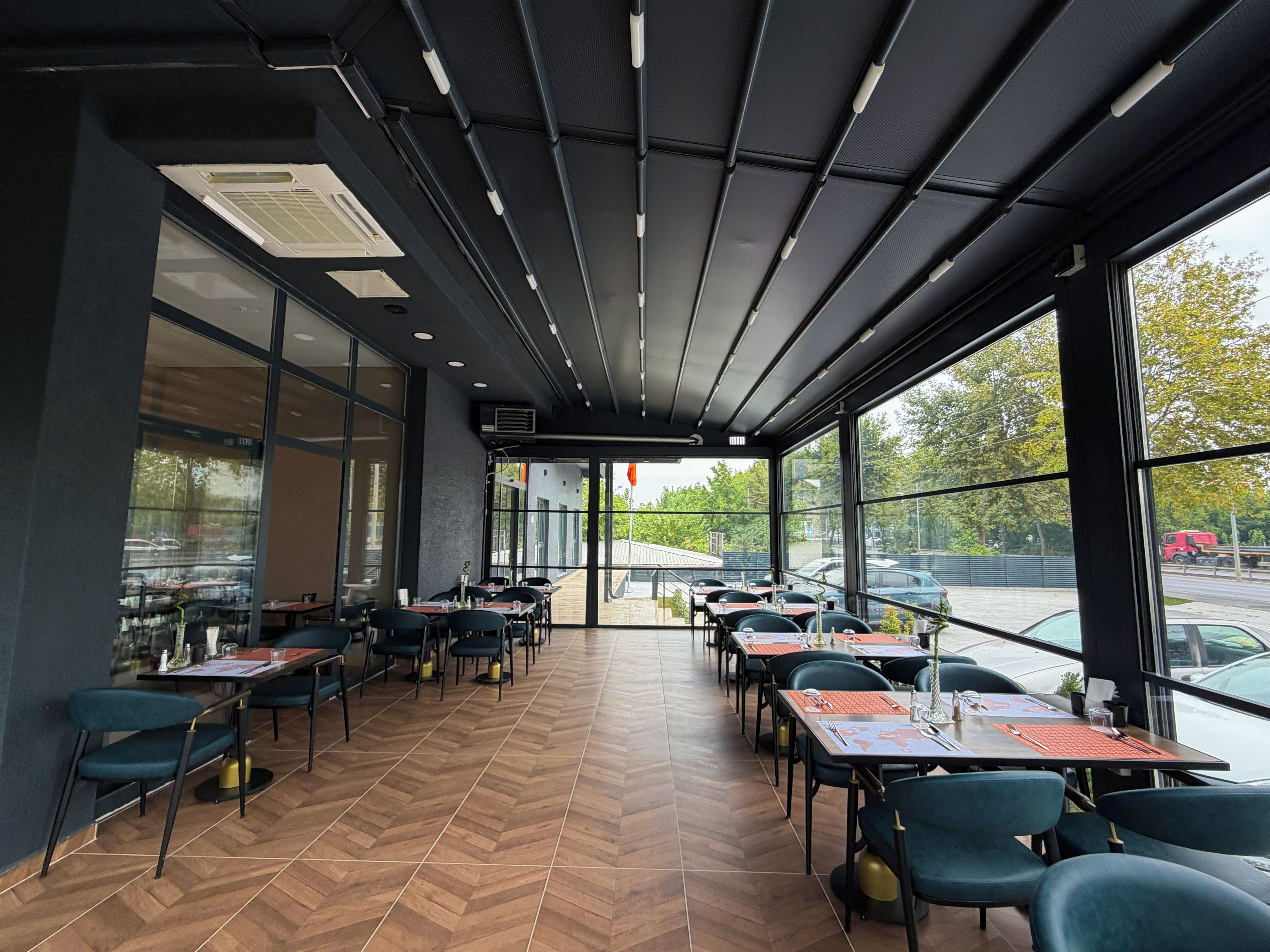 A restaurant with a modern interior, featuring a herringbone floor pattern, black and teal chairs, and large windows offering a view of the parking lot and trees.