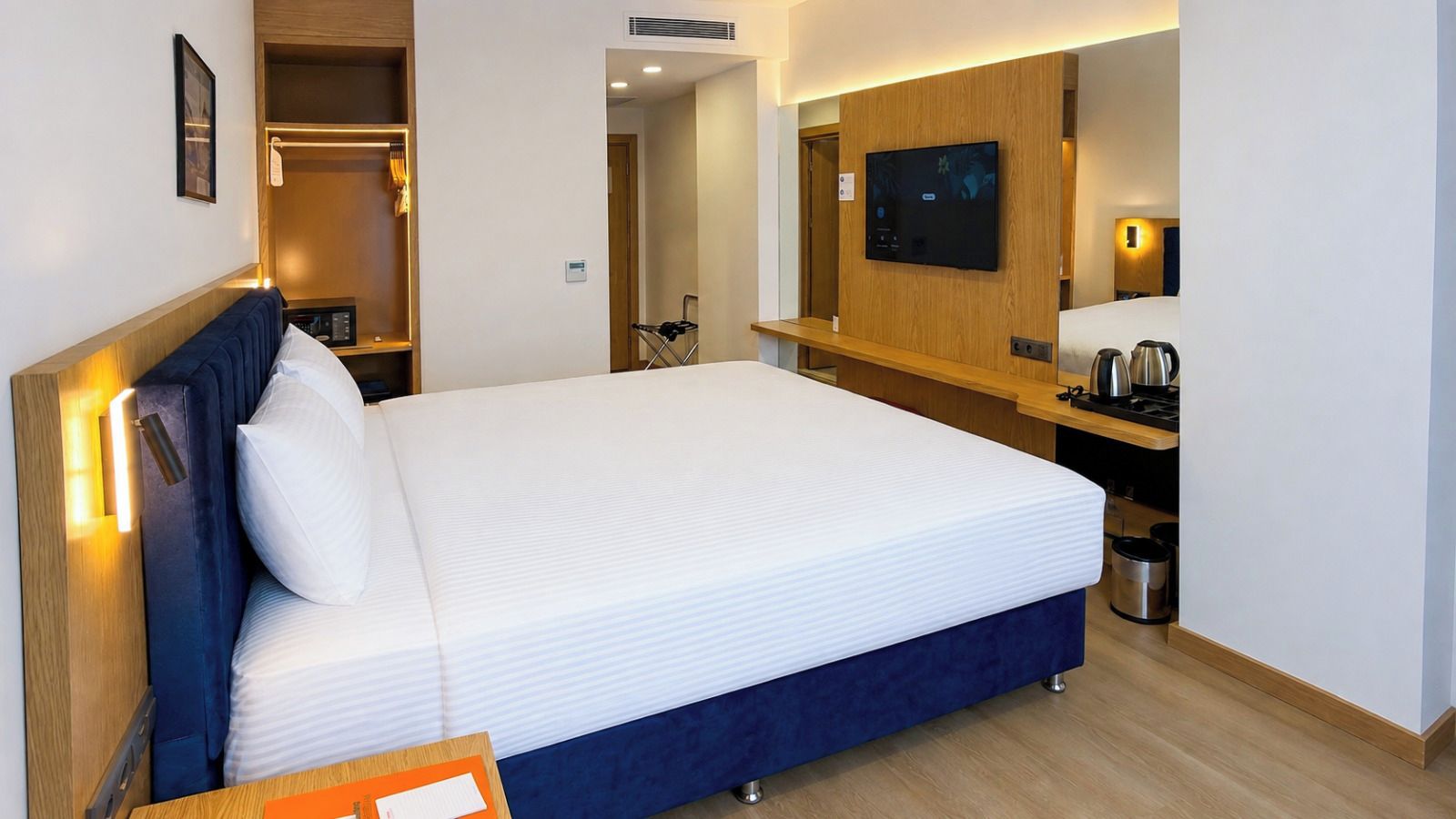A hotel room with a bed, a TV mounted on the wall, a small kitchenette, and a wooden wardrobe.