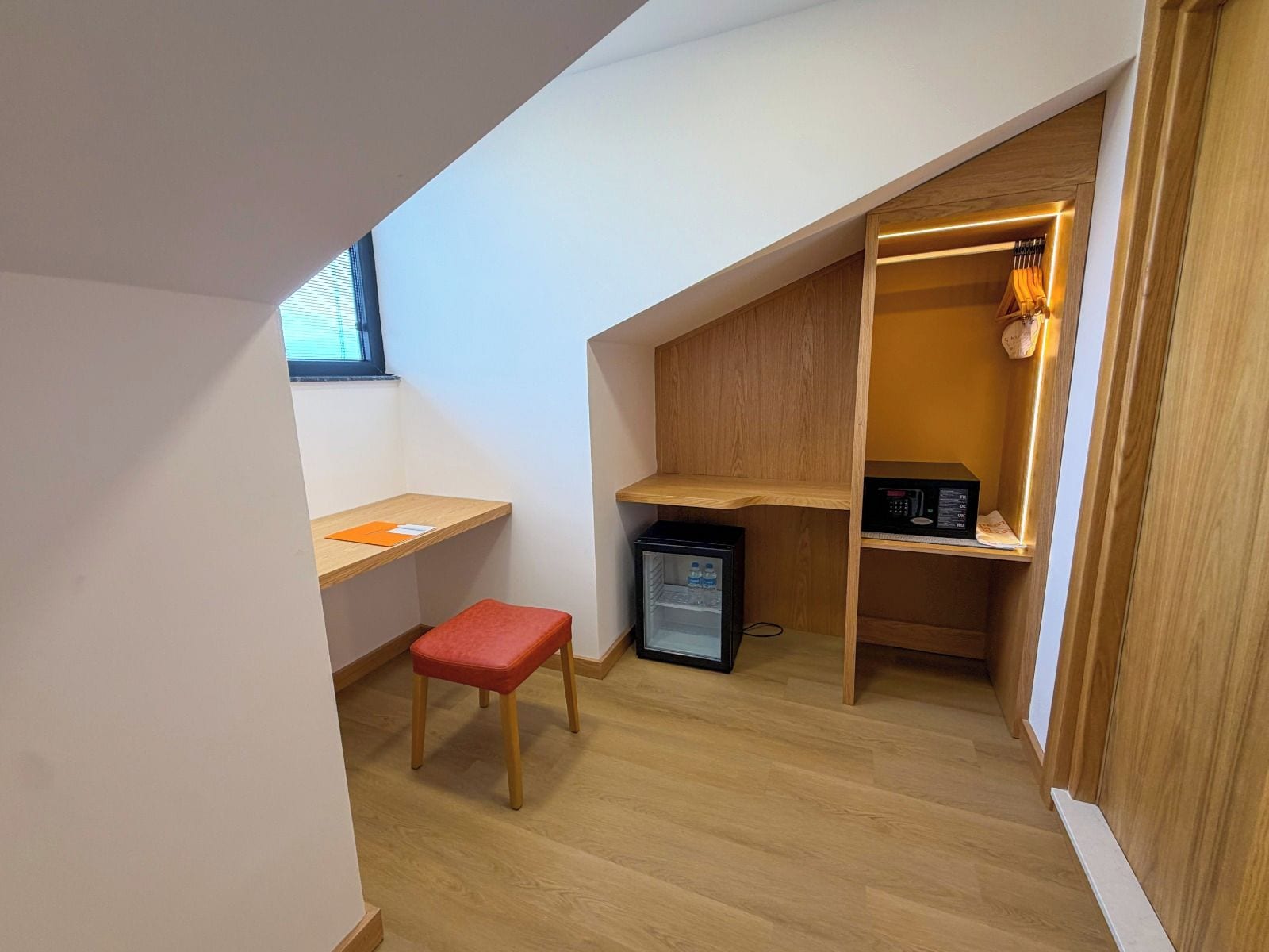 A compact room with a desk, a red chair, a small refrigerator, and a shelving unit with a lamp.