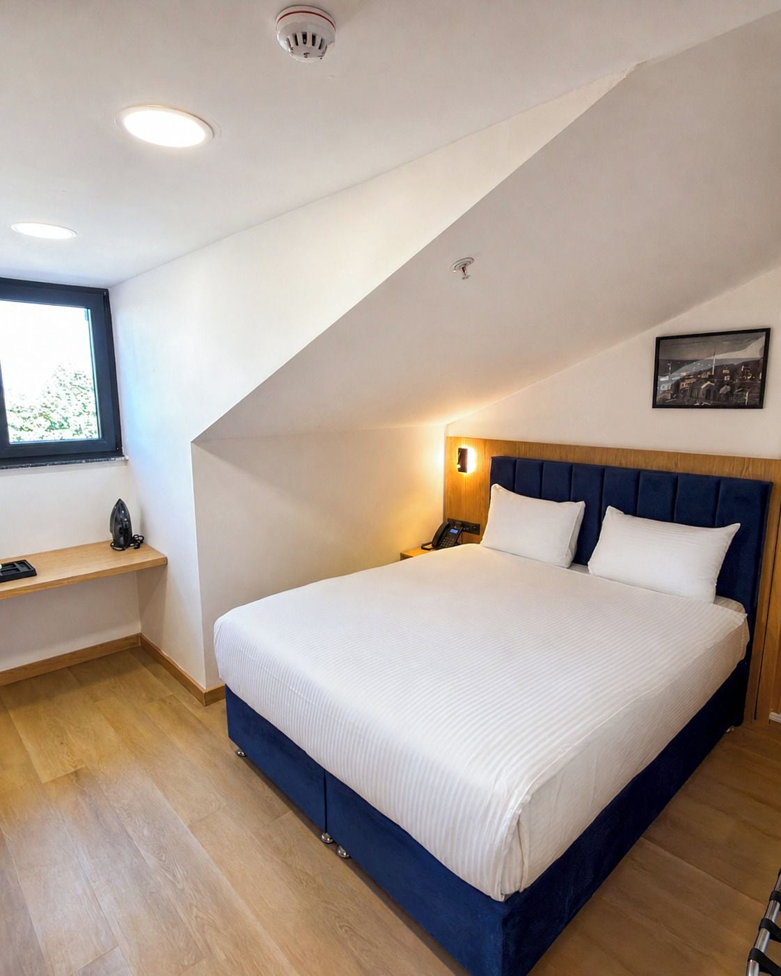 A bedroom with a large bed, a bedside table, a nightstand, a wall-mounted light, a window, and a ceiling-mounted smoke detector.