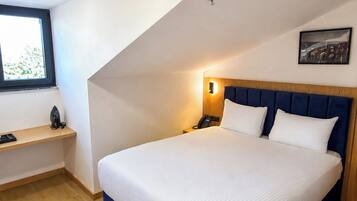 A bedroom with a large bed, a bedside table, a nightstand, a wall-mounted light, a window, and a ceiling-mounted smoke detector.