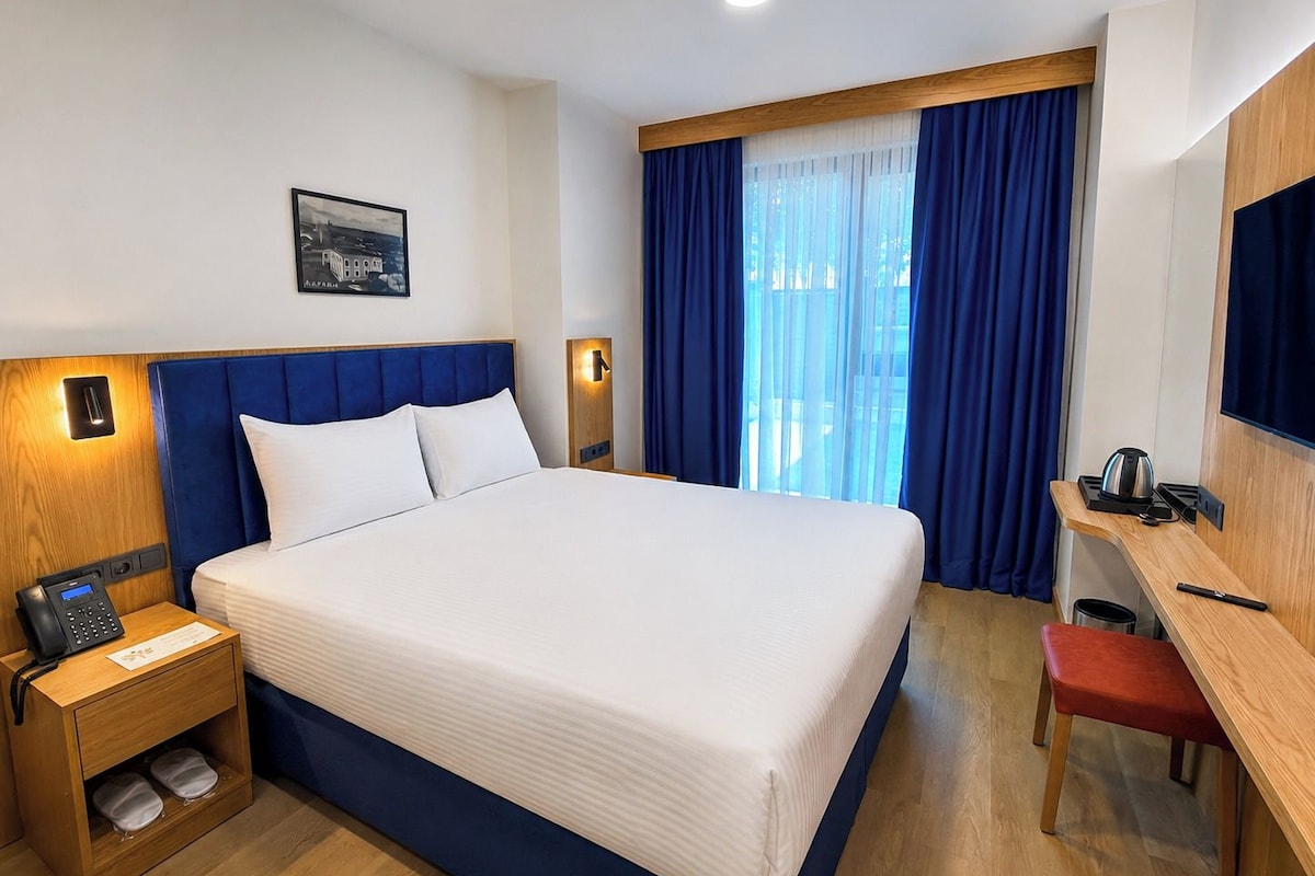 A hotel room with a large bed, a desk with a chair, a small table with a kettle, and a nightstand with a phone.