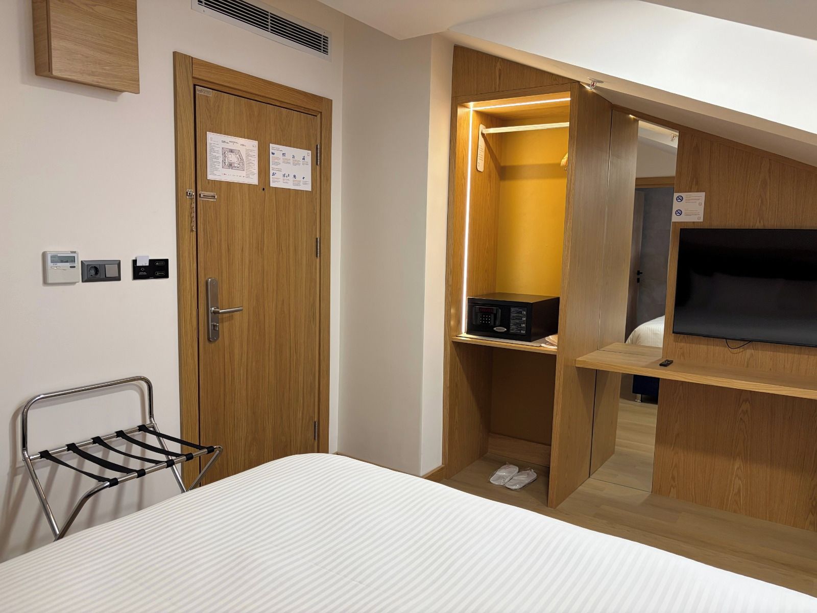 A compact hotel room with a bed, a TV mounted on the wall, a wooden wardrobe, and a shoe rack.