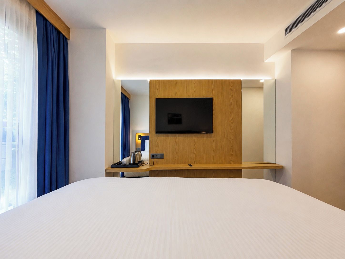 Habitación de hotel con una cama grande, un televisor de pantalla plana montado en un panel de madera, una mesita de noche con hervidor y una ventana con cortinas azules.