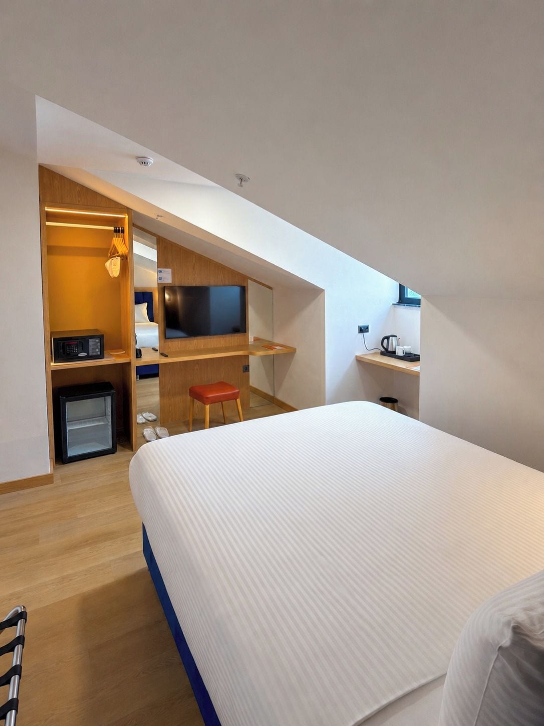 Quarto de hotel moderno com uma cama grande, televisão, escrivaninha com computador e uma pequena cozinha equipada.