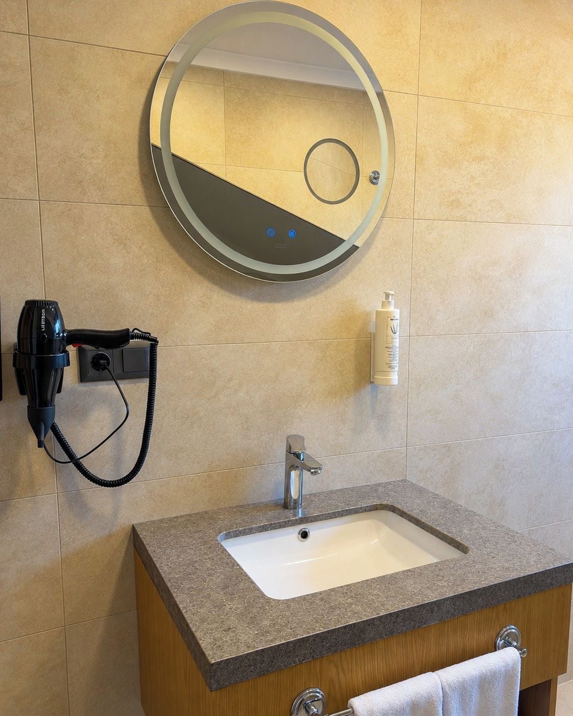 Un baño con un espejo redondo, un lavabo, un secador de pelo y una botella de loción.