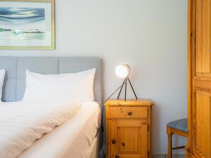 A neatly made bed with white linens, a bedside lamp, and a wooden nightstand with a drawer. - Ground Floor in the Jan Mayen House (St. Peter-Ording)