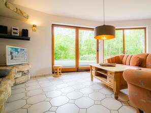 A modern living room with a stone accent wall, a wooden coffee table, a sofa, and a large window with a view of greenery. - Ground Floor in the Jan Mayen House (St. Peter-Ording)