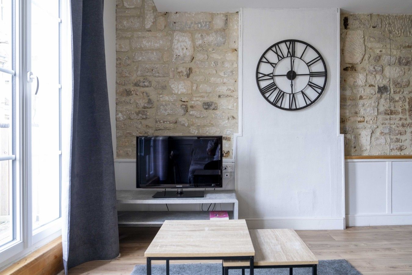 A modern living room with a stone wall, a large black clock, a flat-screen TV, and a minimalist wooden table.