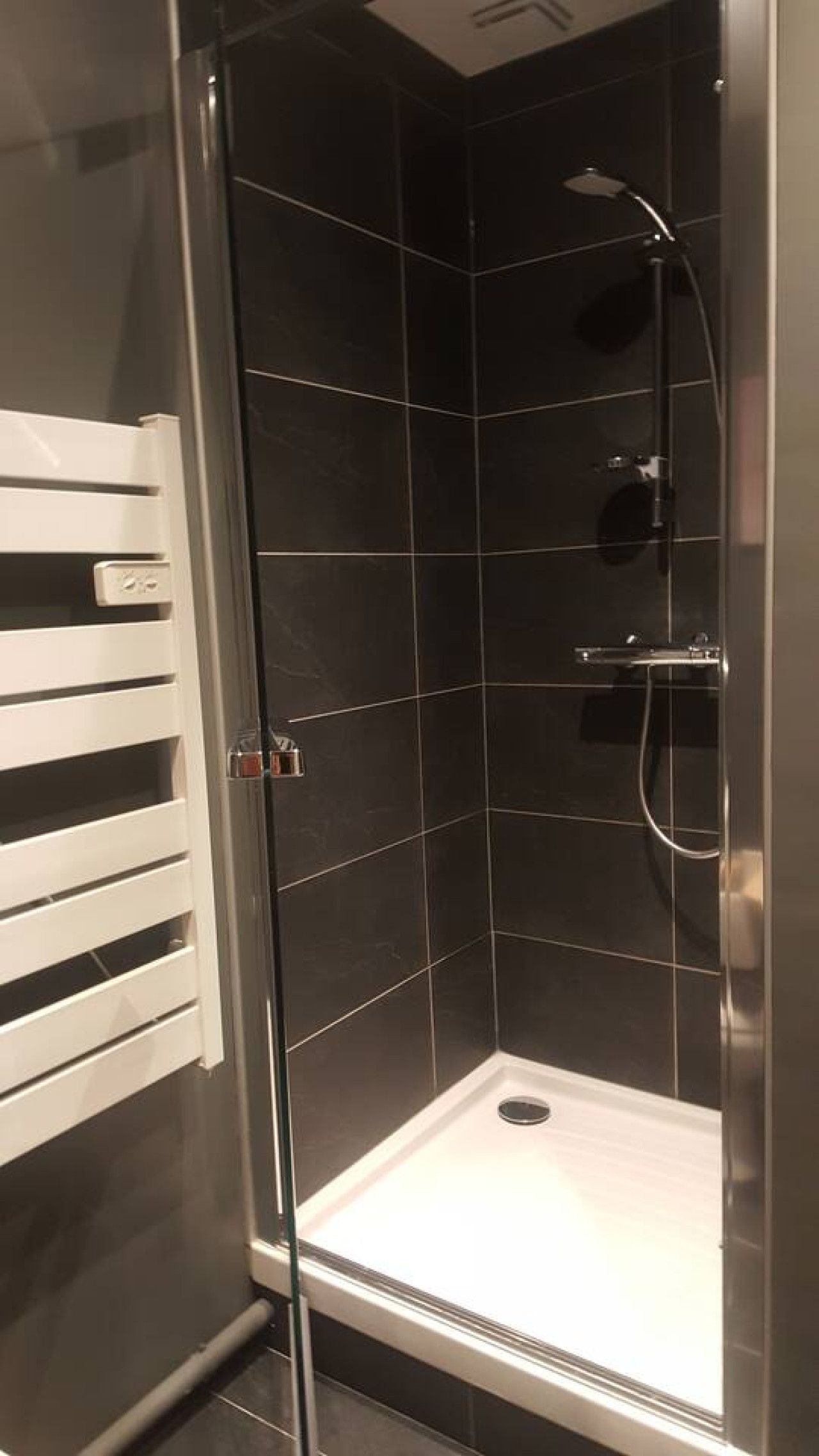 Une cabine de douche fermée par des parois en verre, avec un sol carrelé de blanc, une douchette à main et un robinet fixé au mur.