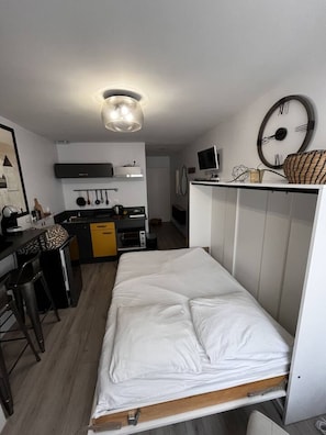 Een kleine kamer met een bed, bureau, kitchenette en garderobe.