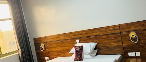 Een hotelkamer met een houten hoofdeinde, een bed met een geborduurd sprei, een nachtkastje met een lamp, en een raam met gordijnen.