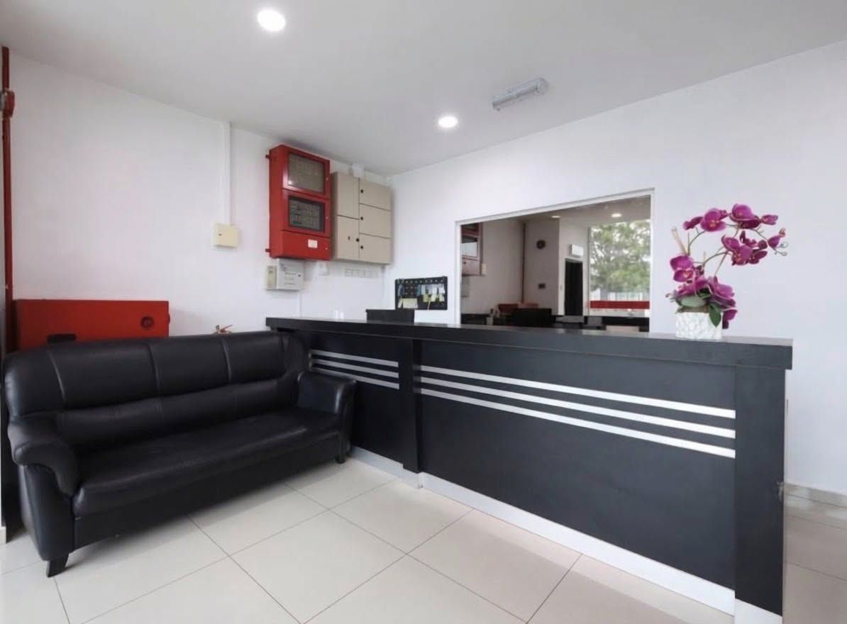 Uma área de recepção com um sofá preto, balcão de atendimento, um extintor de incêndio vermelho e um vaso com flores roxas.