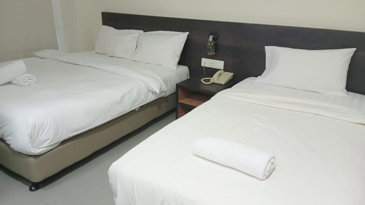 A hotel room with two single beds, each with white linens and pillows, a bedside table with a telephone, and a wall-mounted light fixture.