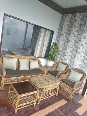 A wicker sofa set with cushions, a small table, and a potted plant in a room with a large window and floral wallpaper.