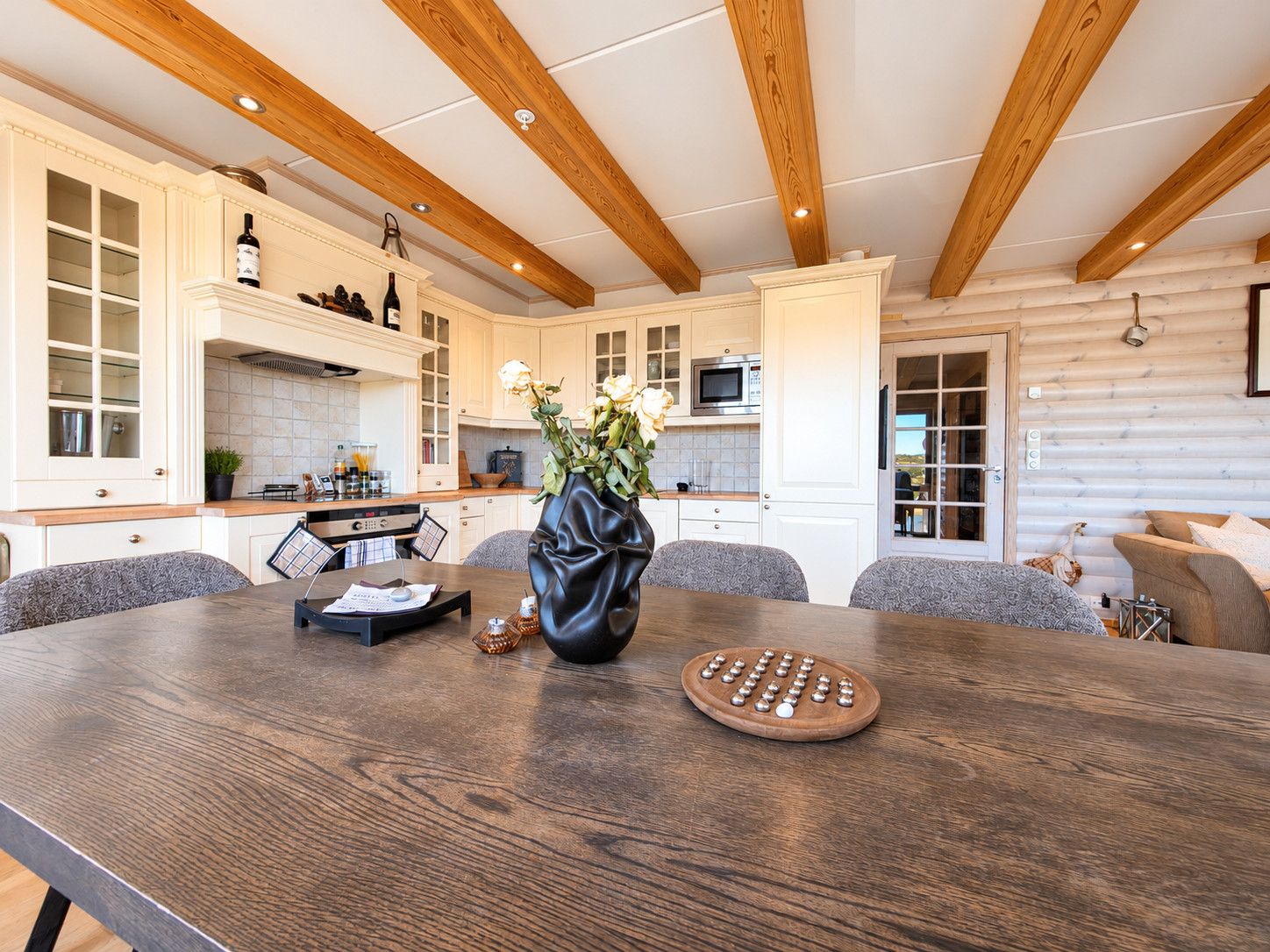 A modern kitchen with wooden beams, a dining table with a vase of flowers, and a wooden board game.