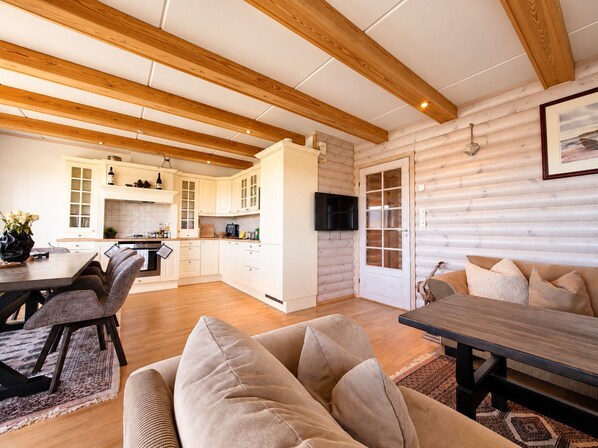Un intérieur moderne avec un plafond en bois, un coin repas avec une table et des chaises, une cuisine avec des meubles blancs et un salon avec un canapé et des coussins.