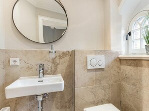 Ein Badezimmer mit einem weißen Waschbecken, einem runden Spiegel, einem an der Wand montierten Lichtschalter und einem Fenster mit einer Pflanze.
