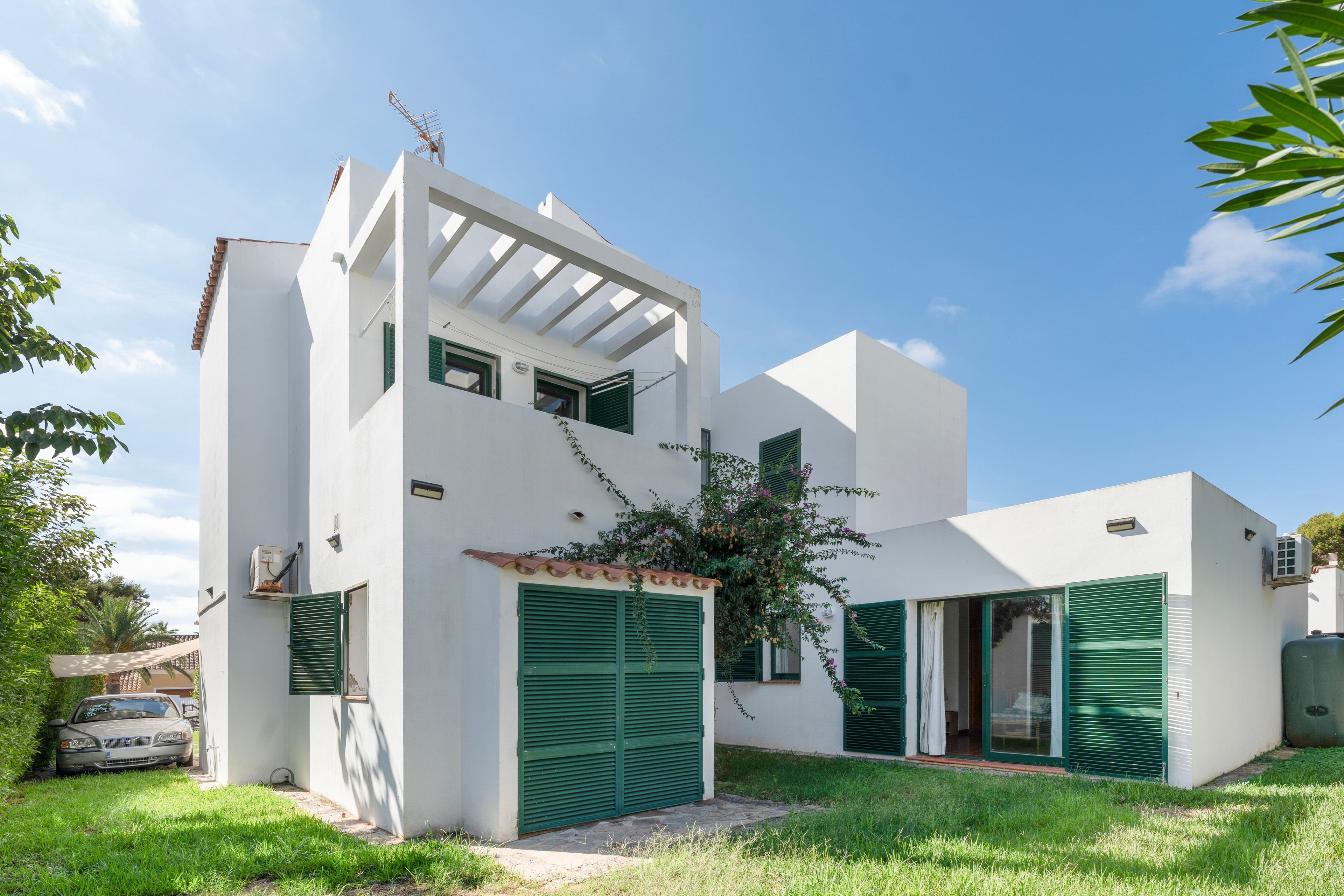 A modern two-story house with a flat roof, green shutters, and a small garden area.