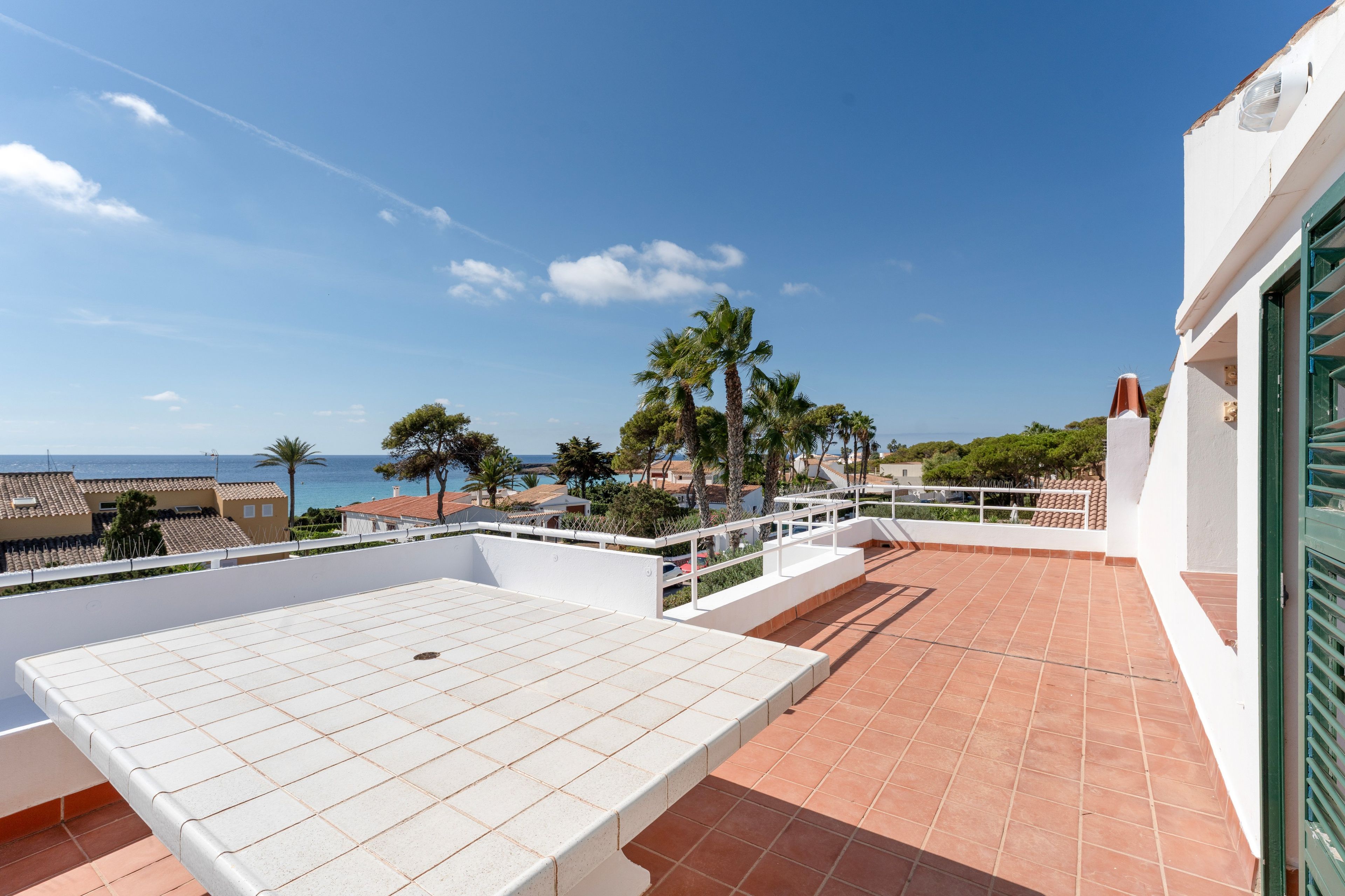 Une terrasse avec une table et des chaises blanches, donnant sur une zone côtière bordée de palmiers et de bâtiments.