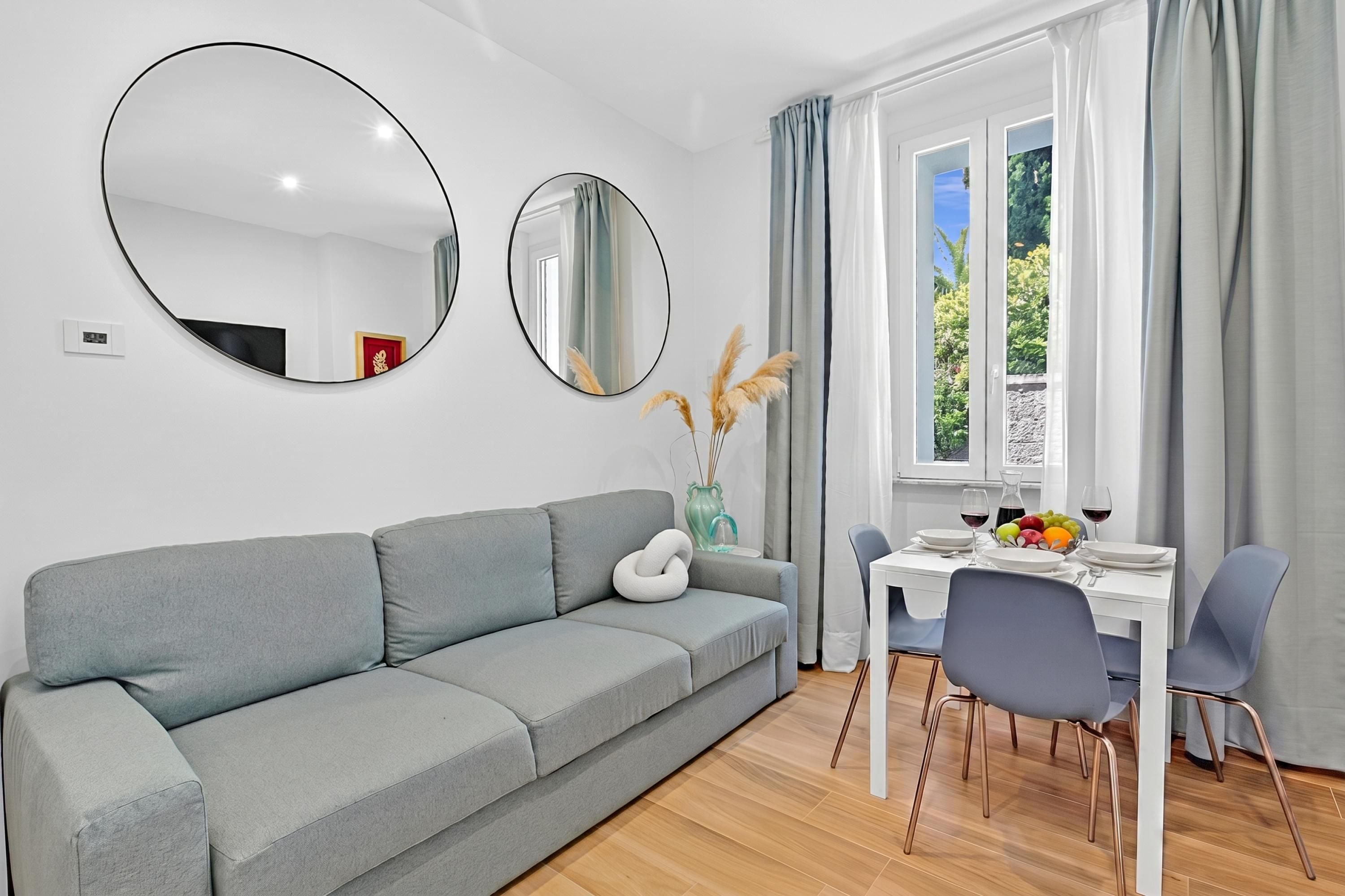 Ein modernes Wohnzimmer mit einem grauen Sofa, einem Esstisch mit Stühlen und zwei runden Spiegeln an der Wand.