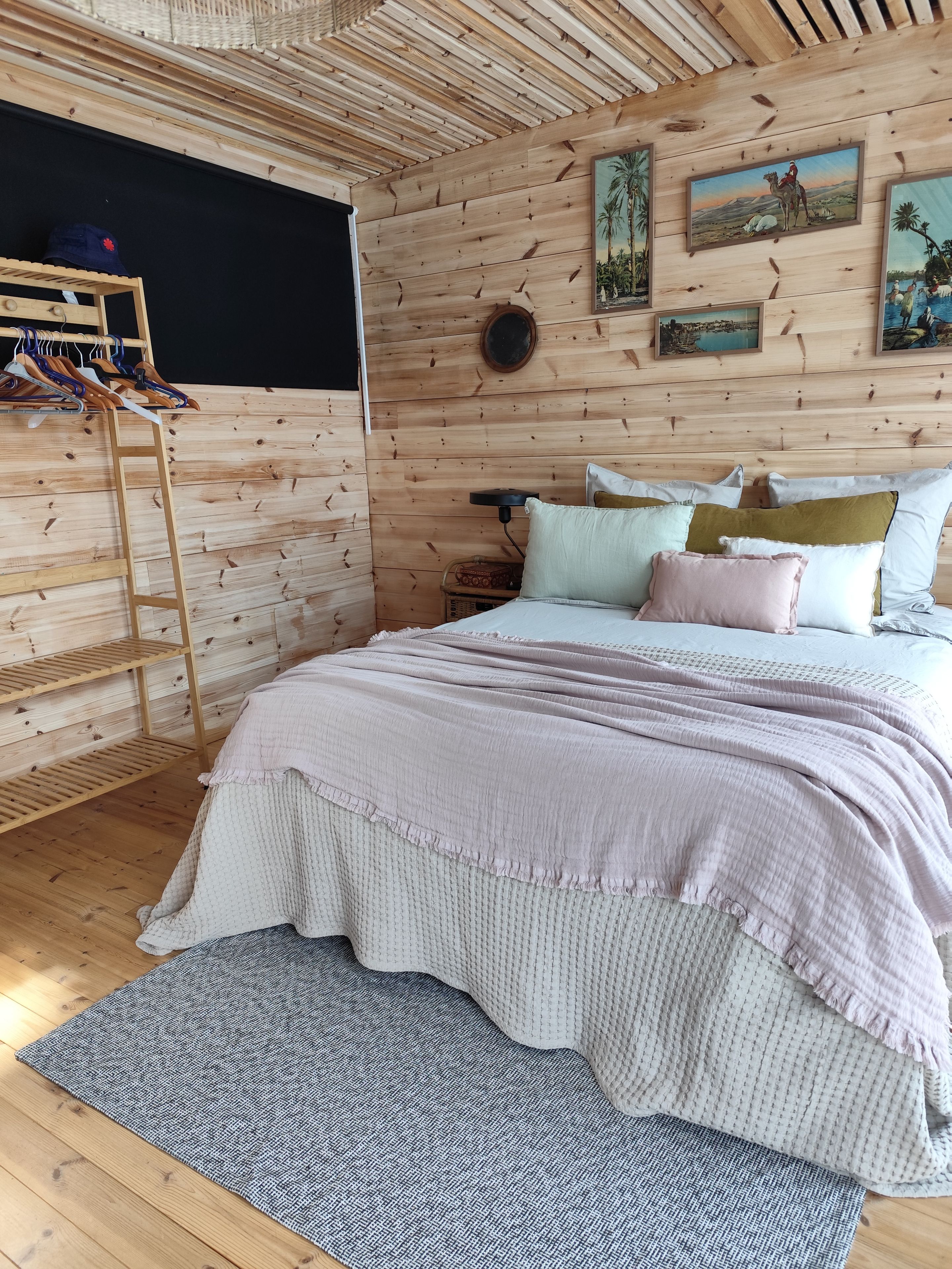 Une chambre avec un plafond en lambris de bois, un lit recouvert de plusieurs coussins, une étagère fixée au mur où sont pliés des vêtements, et une petite table ronde.