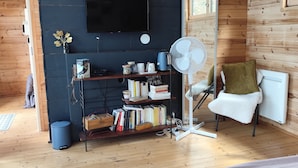 A modern interior with a flat-screen TV mounted on a dark wall, a bookshelf with books and decorative items, a standing fan, and a cozy seating area with a chair and cushions. - Peaceful 1-bedroom bed & breakfast in charming Chantraine (Chantraine)