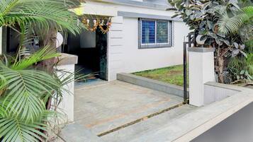 A residential entrance with a concrete pathway leading to a door, surrounded by greenery and a small garden.