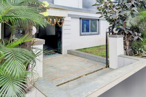 A residential entrance with a concrete pathway leading to a door, surrounded by greenery and a small garden.