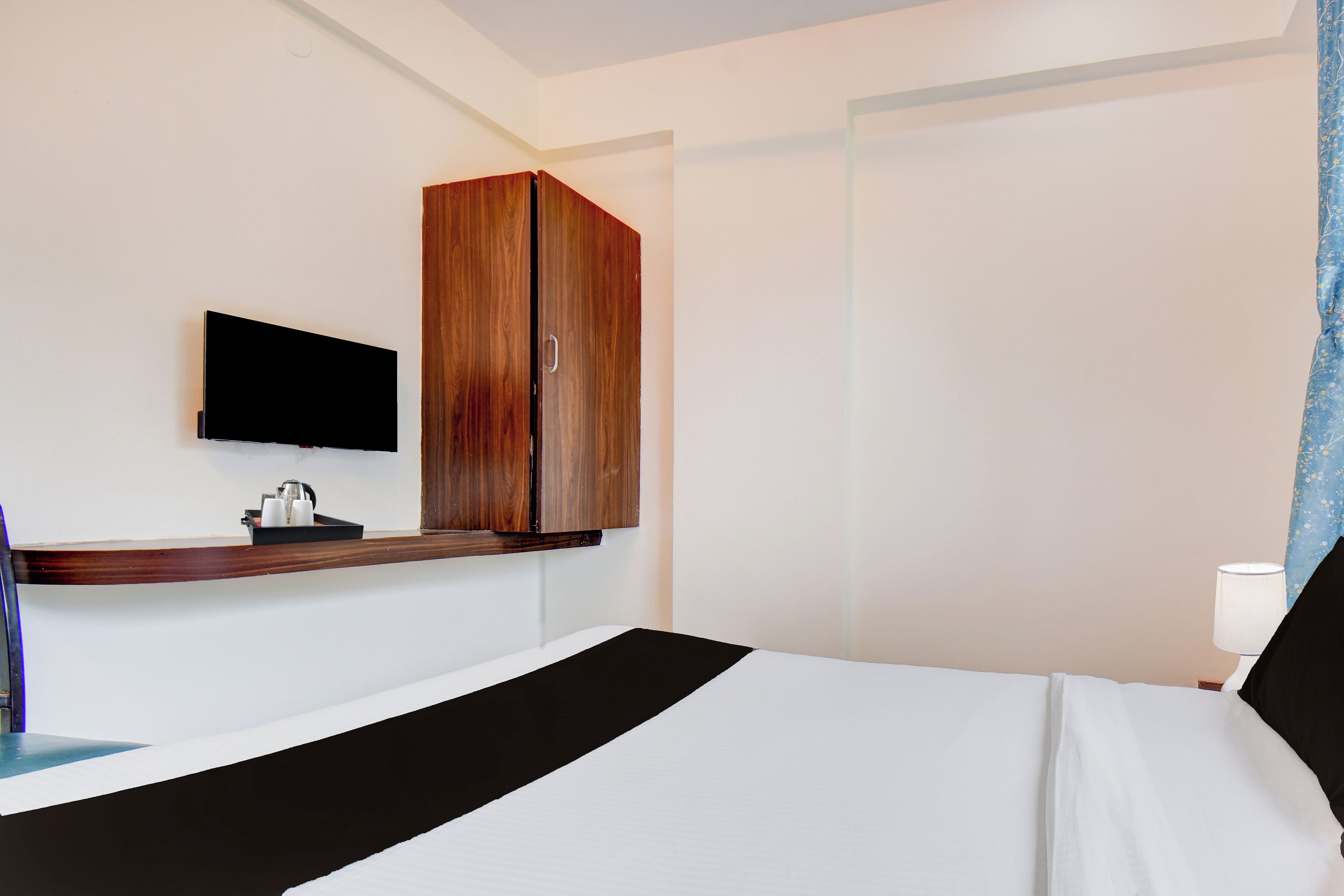 A hotel room with a bed, a television mounted on the wall, a wooden wardrobe, and a bedside table with a tray of items.