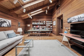 Ein gemütliches Wohnzimmer mit einer Couch, einem Couchtisch und einem Tischtennisschrank. Bücherregale voller Bücher und ein Fernseher sind auf der rechten Seite zu sehen.
