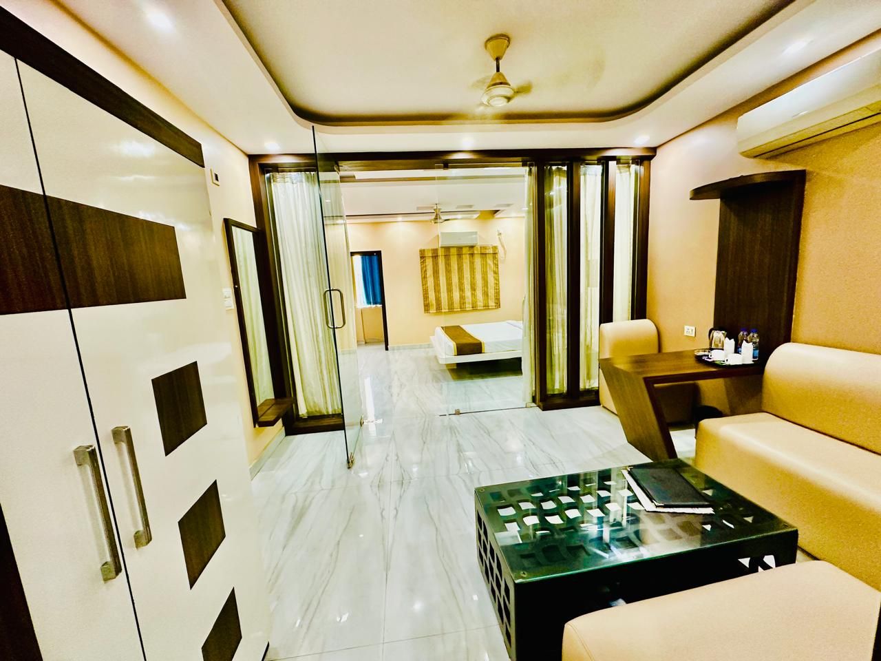 A modern hotel room with a glass partition, a bed, a sofa, a coffee table, and a desk with a lamp.