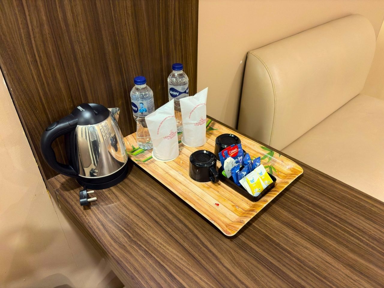 Una bandeja de madera con una tetera, botellas de agua, una cafetera y aperitivos envasados sobre una mesa en una habitación con un sofá color beige.