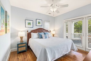 A bedroom with a bed, two bedside tables with lamps, a ceiling fan, and a large window with blinds. - It's 5 O'Clock Somewhere! Ocean Views, Steps to the Beach by Tybee Cottages (Tybee Island)