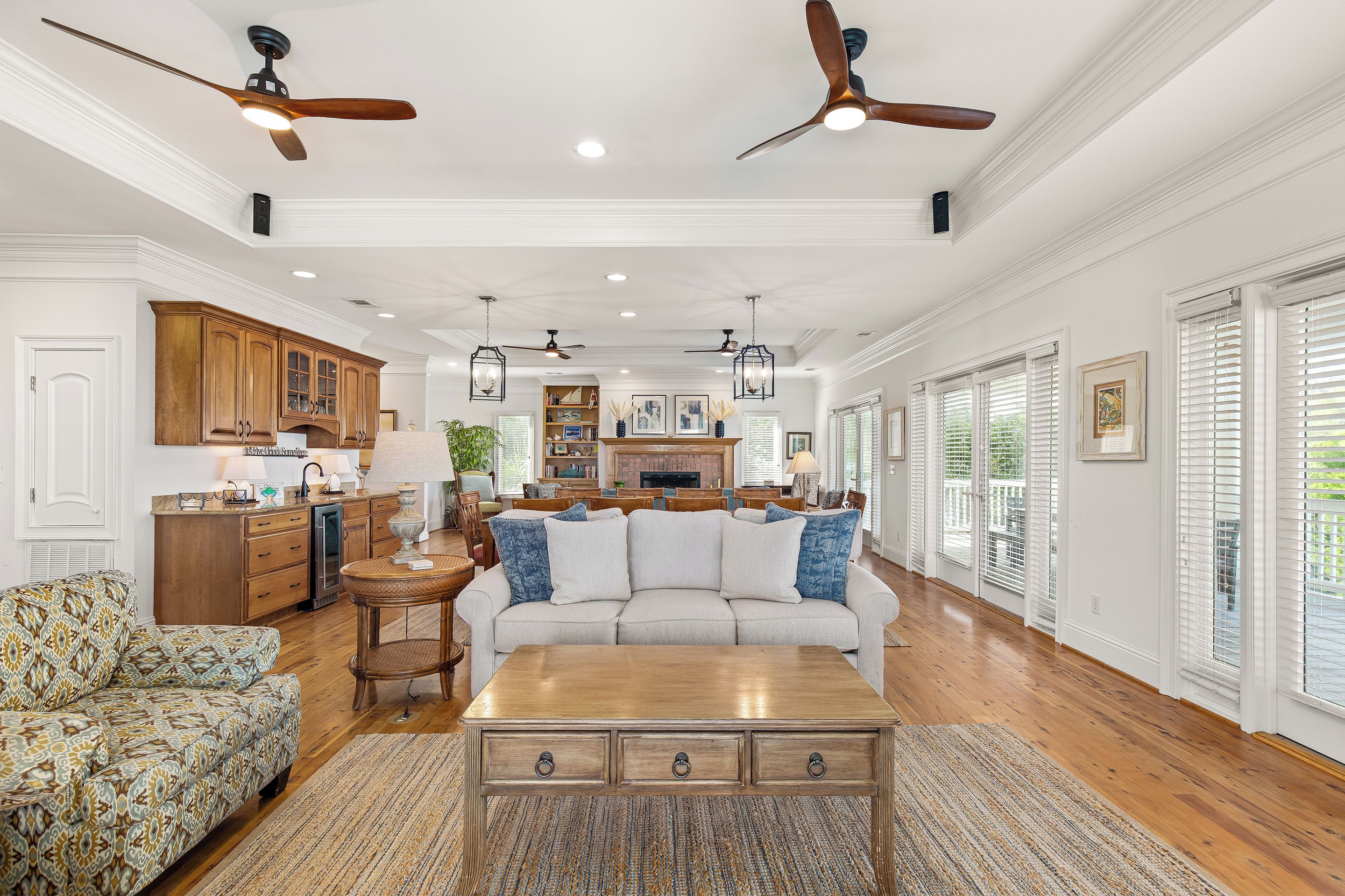 A spacious living room with a large sofa, a coffee table, and a fireplace. There are ceiling fans, recessed lighting, and a view of the outdoors through large windows.