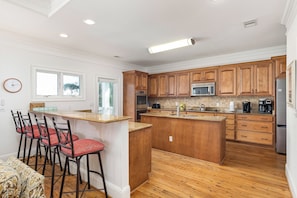 A kitchen with wooden cabinets, a central island, and a dining area with red chairs. - It's 5 O'Clock Somewhere! Ocean Views, Steps to the Beach by Tybee Cottages (Tybee Island)
