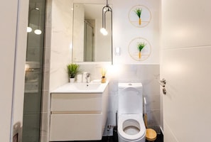 Un baño moderno con un lavamanos blanco, inodoro y un espejo sobre el lavamanos. Hay dos plantas colgantes en la pared y una lámpara colgante.