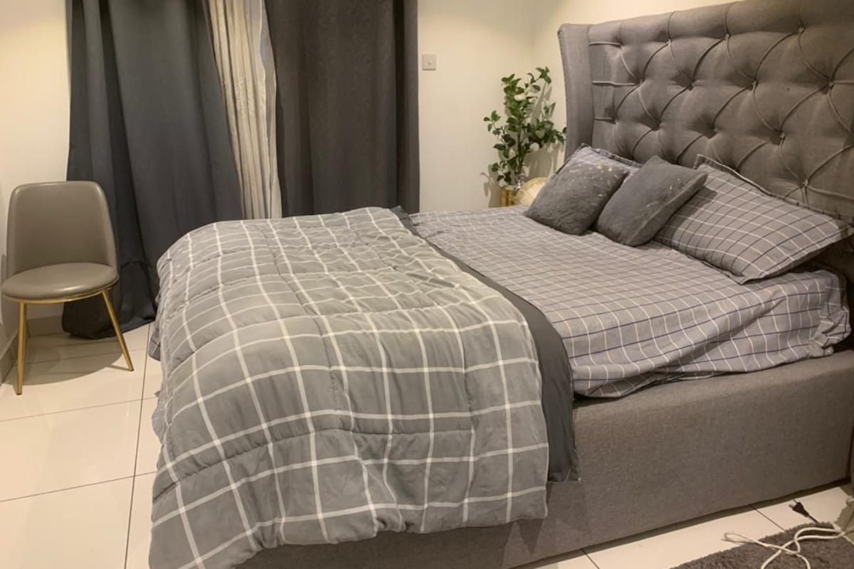 Un dormitorio con cabecera capitonada, colcha a cuadros grises y una silla tapizada en gris.
