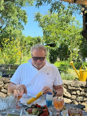 A person seated at an outdoor table with a meal, surrounded by greenery and a stone wall.