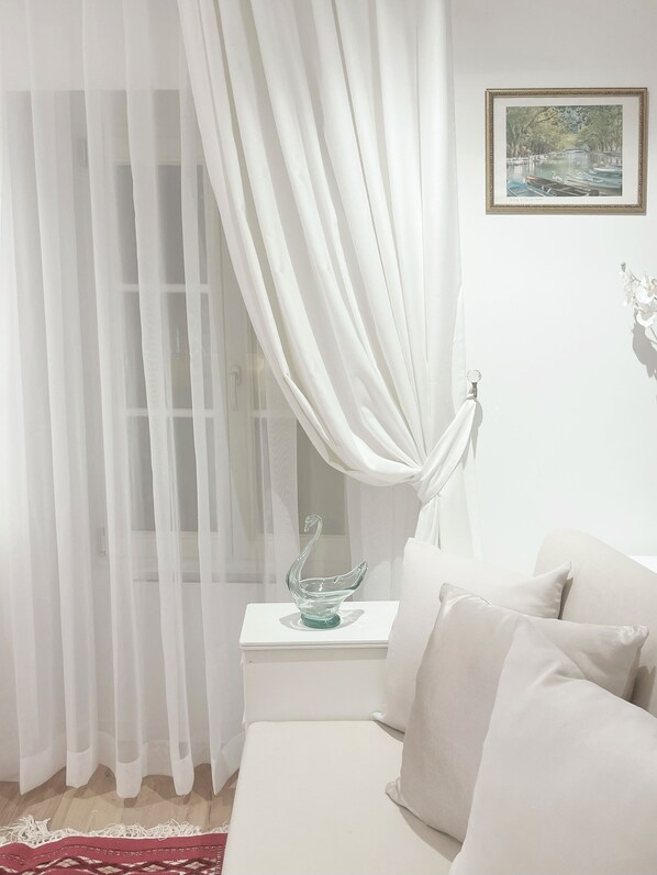 A room with a bed, a nightstand, a glass swan decoration, sheer white curtains, and a framed picture on the wall.