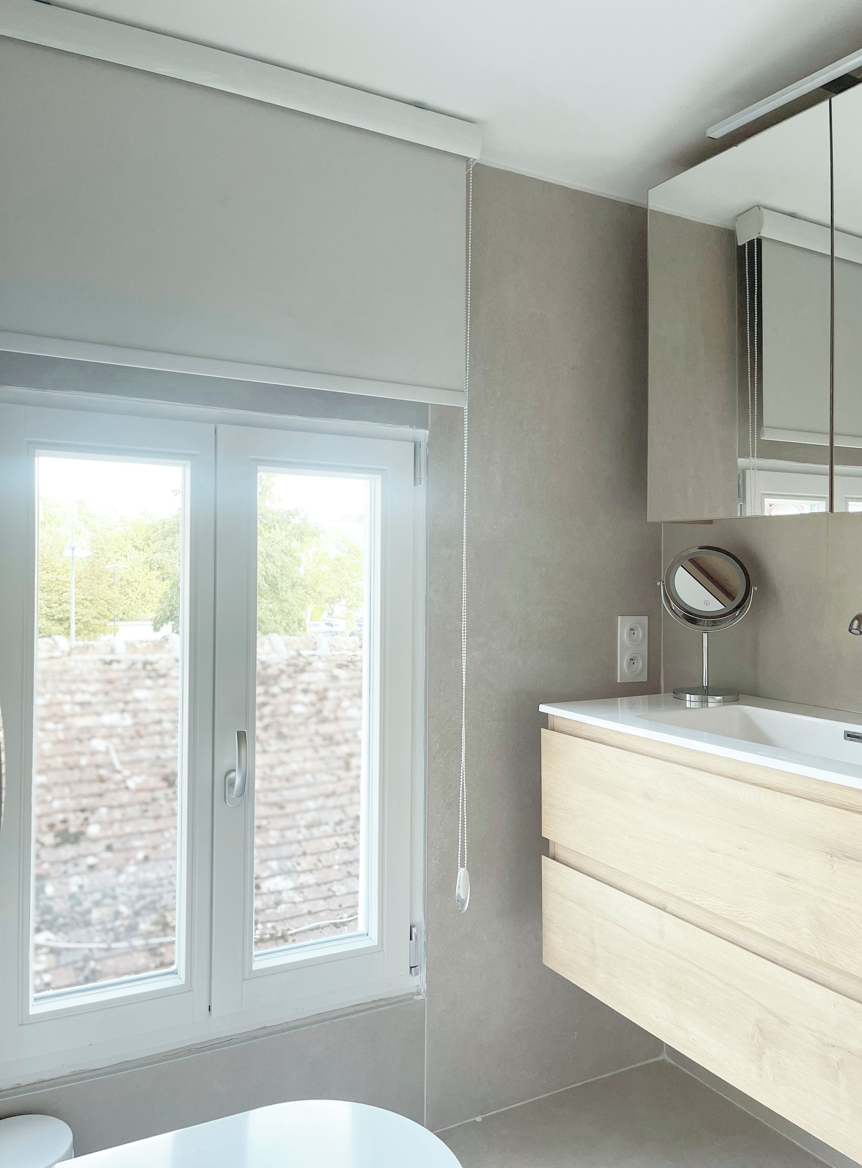 A modern bathroom with a large window, a round mirror, and a wooden vanity.