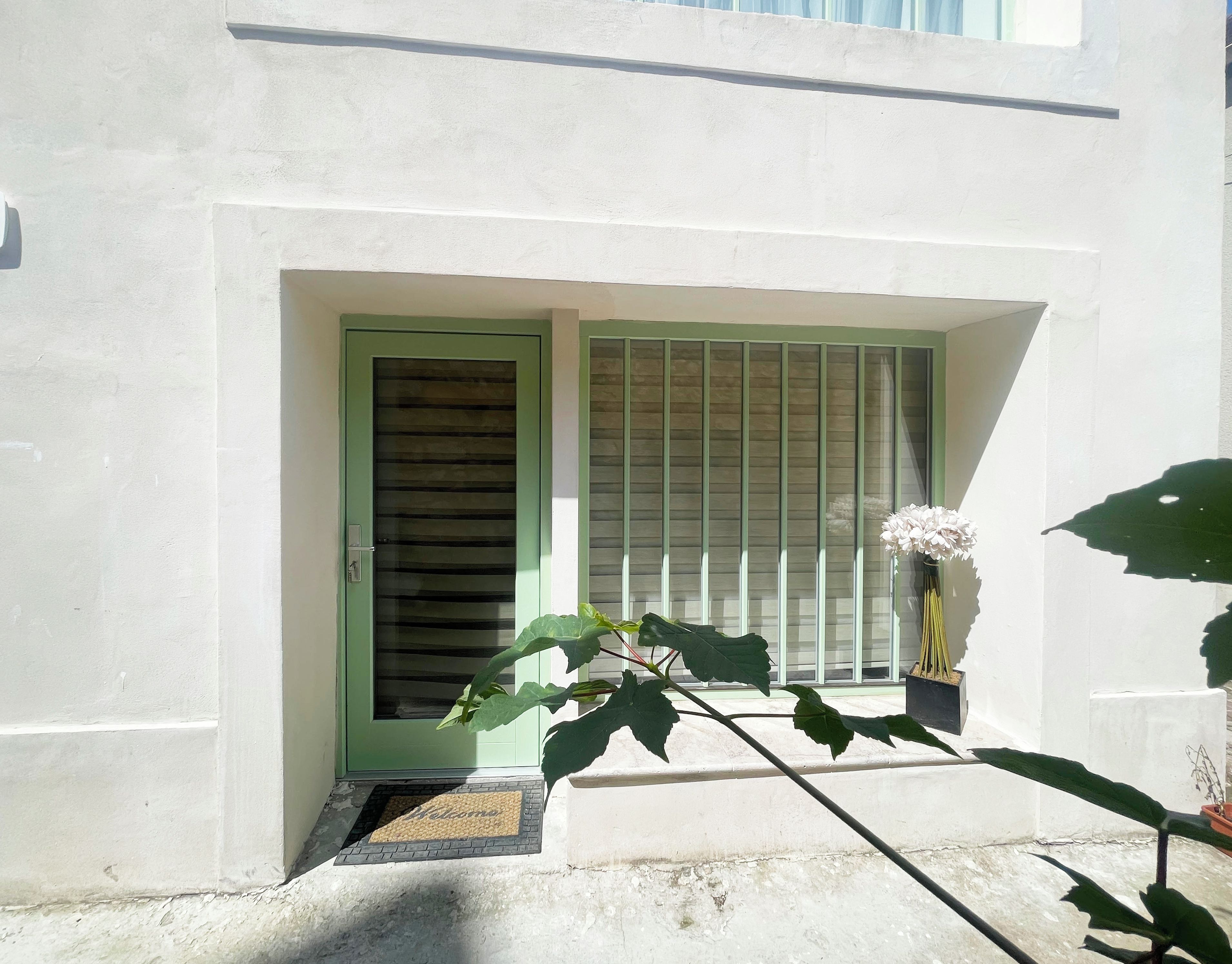 Una entrada de edificio con una puerta verde, una ventana con persianas y una planta en maceta con flores blancas.