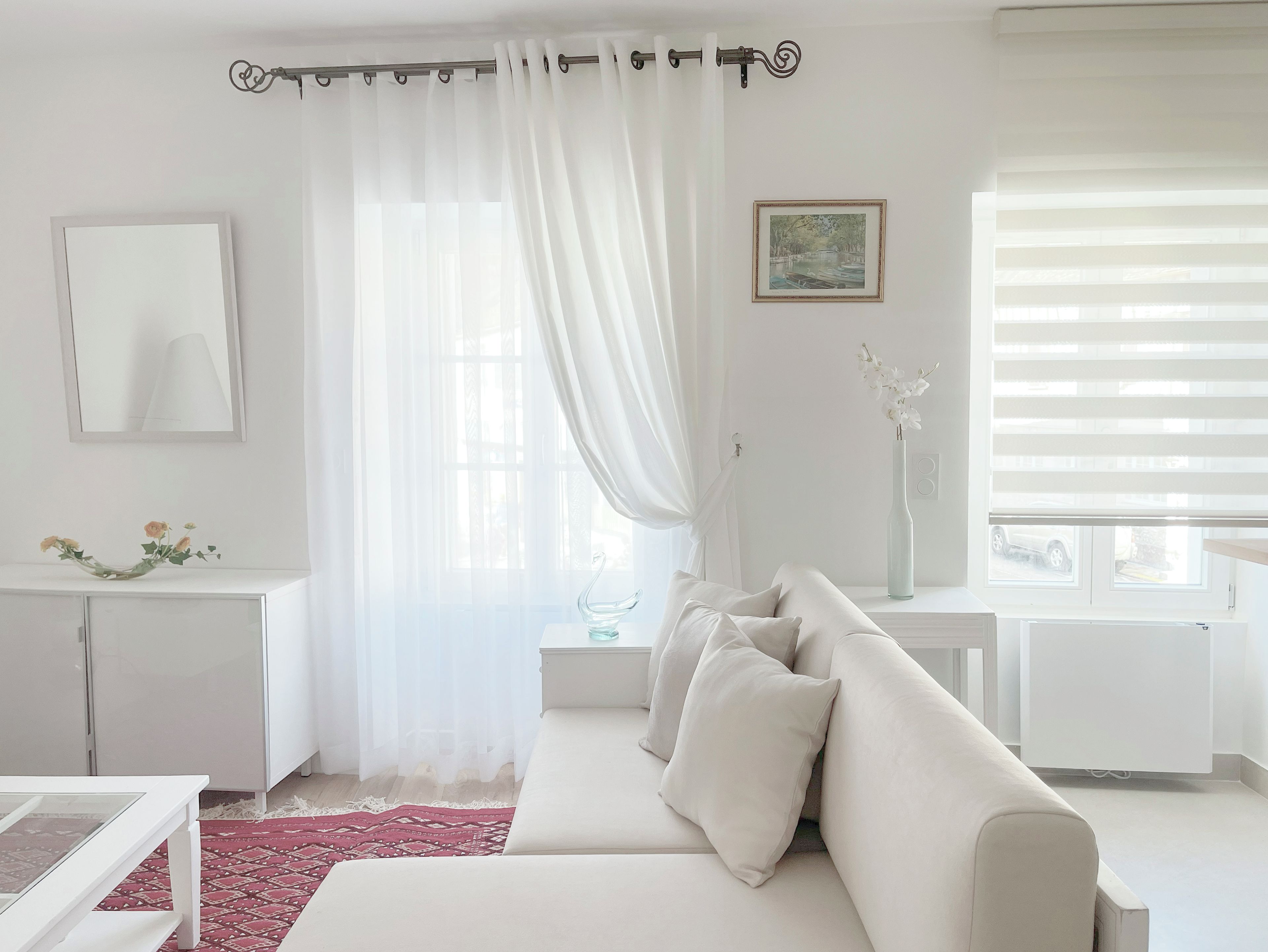 Un salon moderne avec un canapé blanc, une table basse en verre, un tapis à motifs rouges, une console blanche ornée d’un vase de fleurs et une fenêtre avec des rideaux légers.