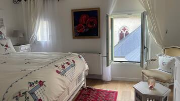 Een slaapkamer met een bed, een raam met gordijnen, een schilderij en uitzicht op een gebouw.