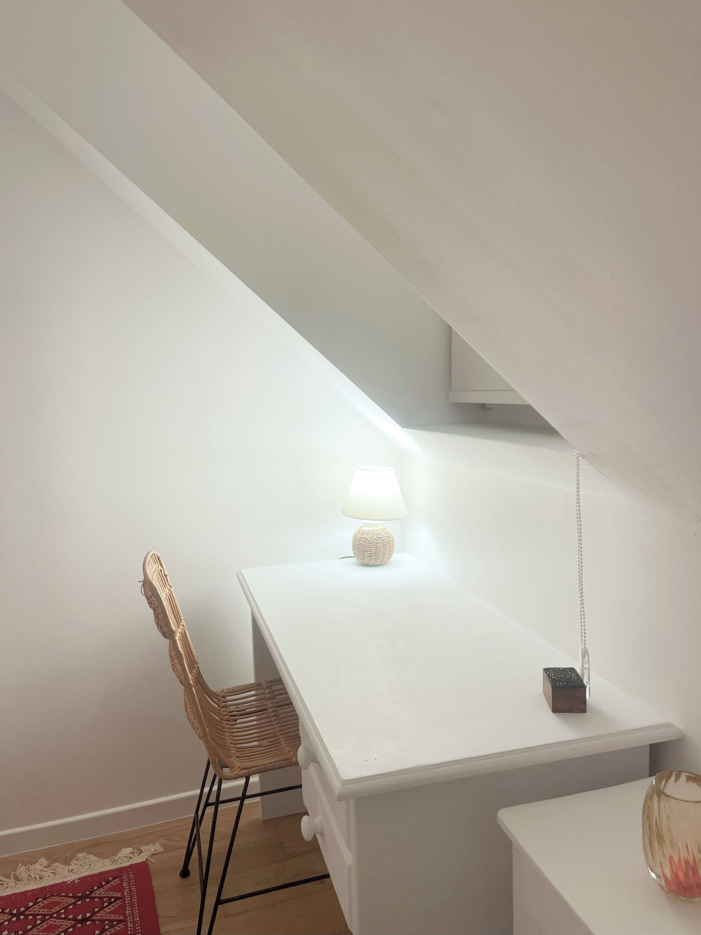 A white desk with a lamp, a wicker chair, and a small table with a decorative item.