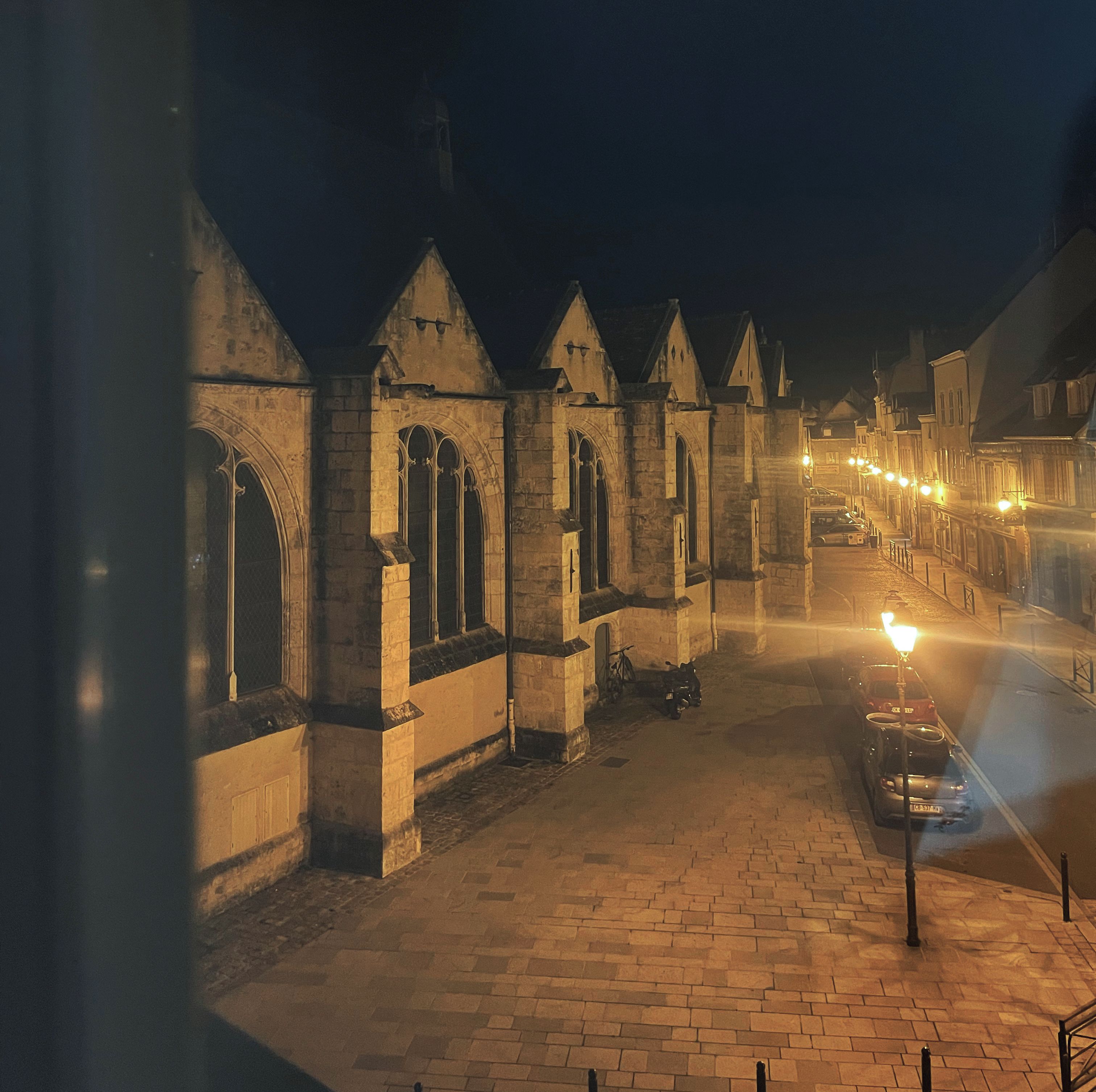 Une vue nocturne d’un bâtiment historique en pierre, doté d’arcs en plein cintre au niveau des fenêtres et situé sur une rue pavée.
