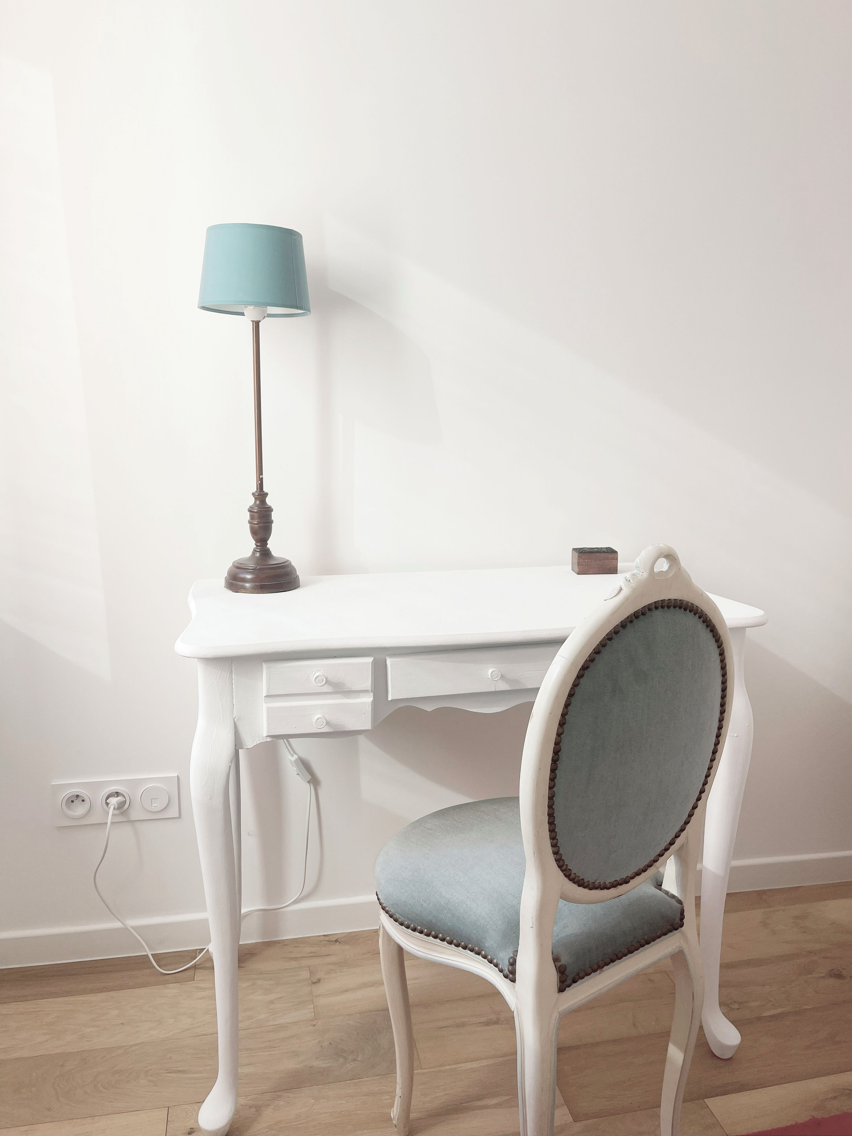A white desk with a lamp, a chair with a cushioned seat, and a power outlet on the wall.