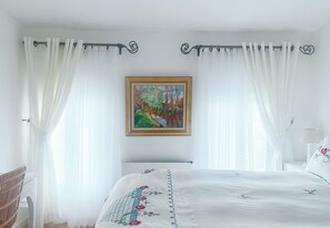 A bedroom with a bed, white curtains, a painting on the wall, and a lamp.