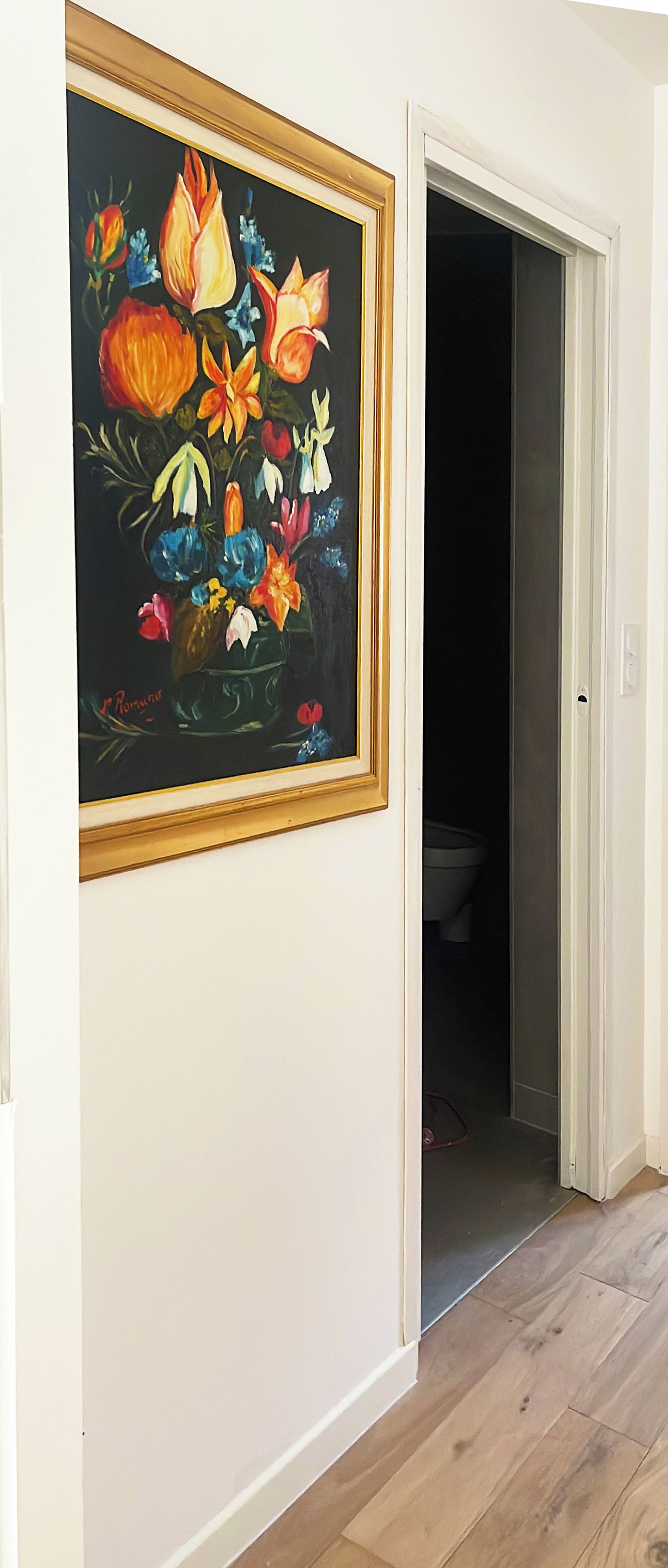 Een gang met een lijstwerk schilderij van bloemen aan de muur en een deur die naar een donkere kamer leidt.